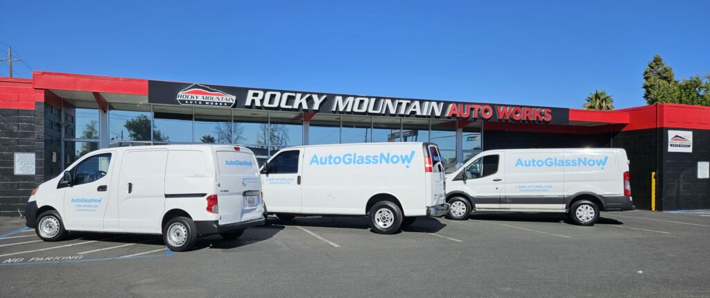 Three white vans with blue AutoGlassNow! decals on them in front of Rocky Mountain Auto Works.