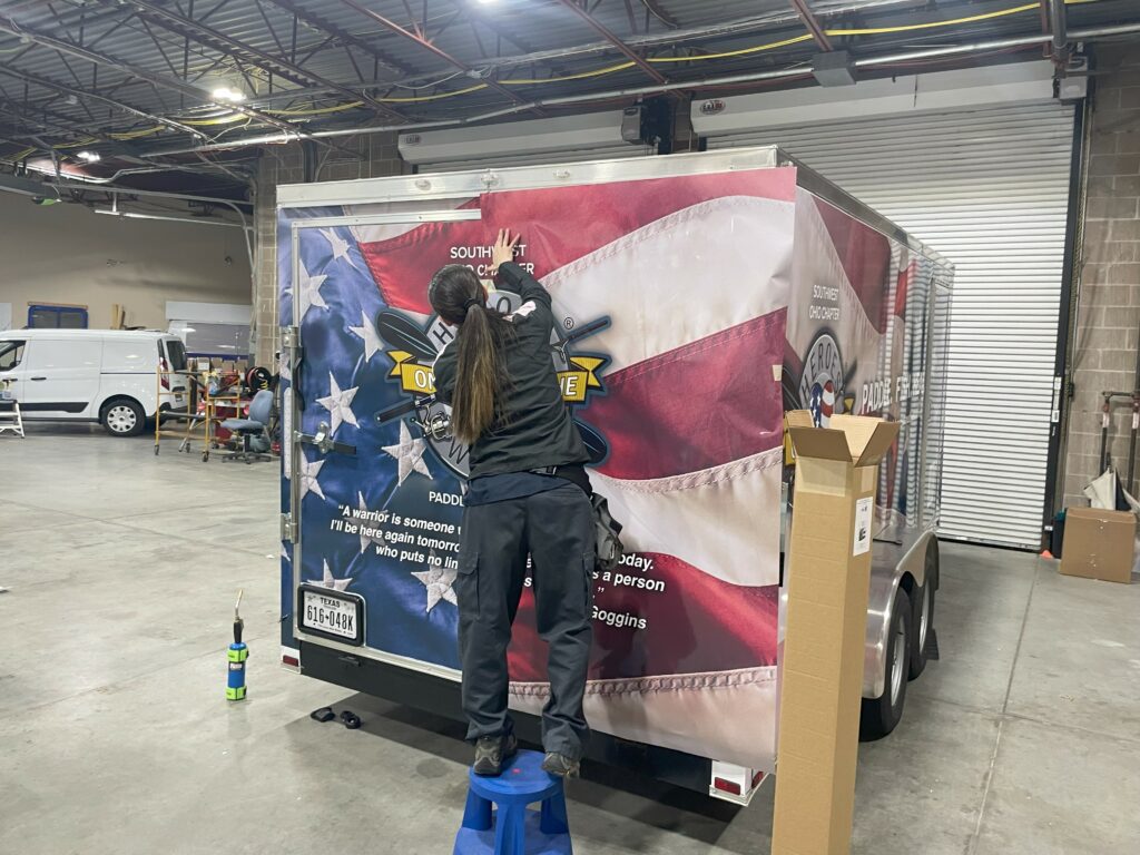 Photo of a woman applying the vinyl graphics onto a trailer. The graphic is of the American Flag with the Heroes on the Water logo.